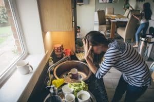 Stressed mum at home. She has her head in her hands at a messy kitchen sink and her children are running round in the background.