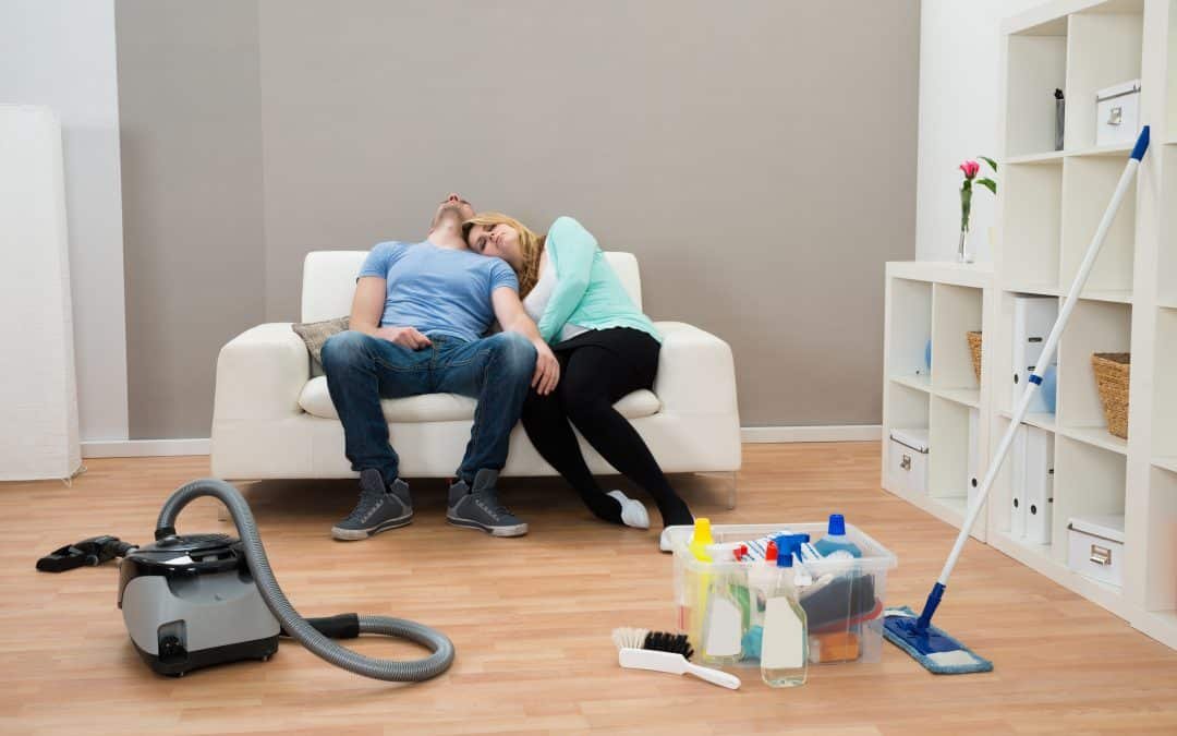 Exhausted Couple Resting On Sofa In Living Room with cleaning supplies abandoned